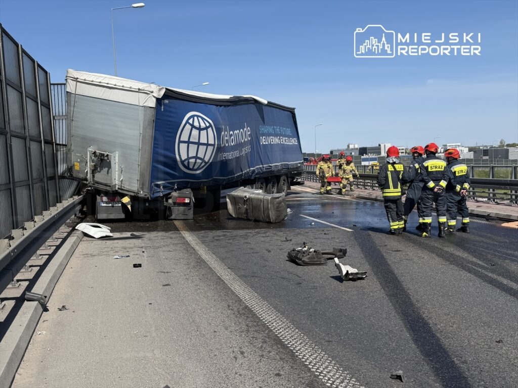 Czterech strażaków w kaskach obserwujących wypadek na drodze, obok przewrócony tir z uszkodzoną naczepą i odłamkami na jezdni.