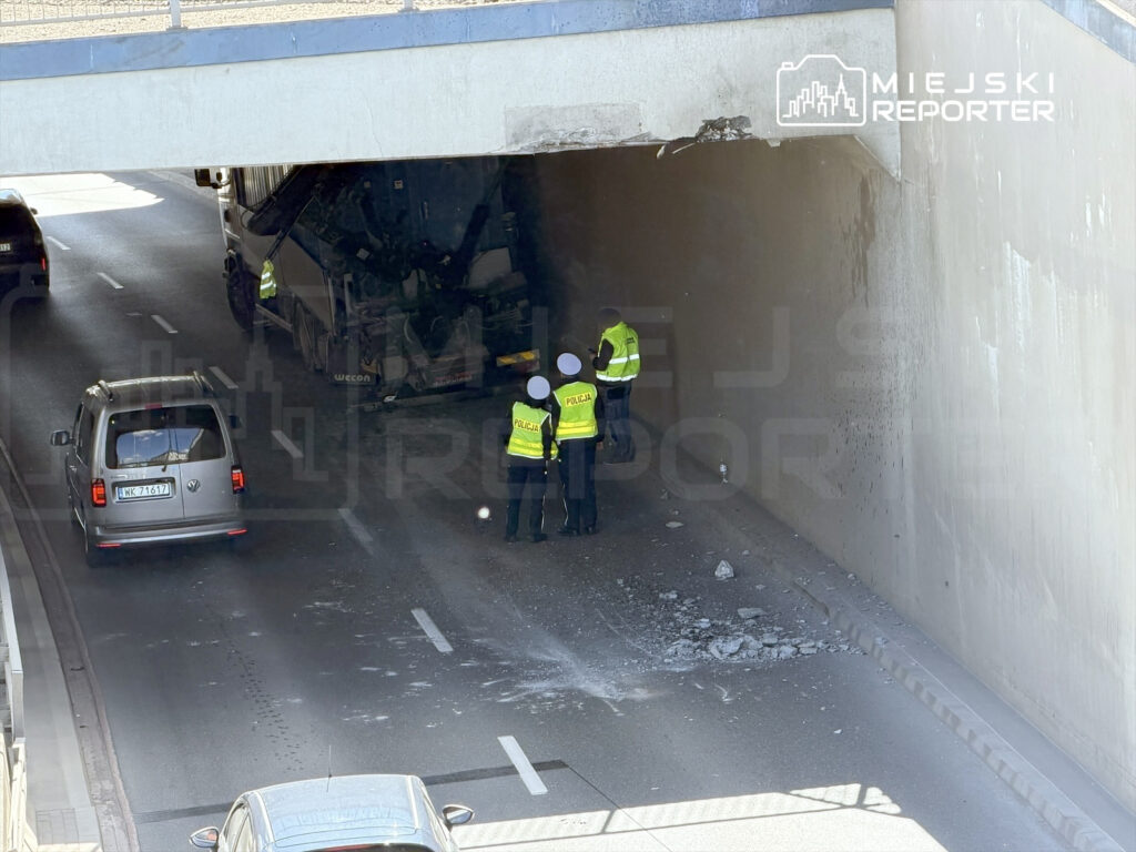 Policjanci w kamizelkach odblaskowych badają uszkodzenia pod wiaduktem drogowym, obok przewróconego pojazdu ciężarowego.