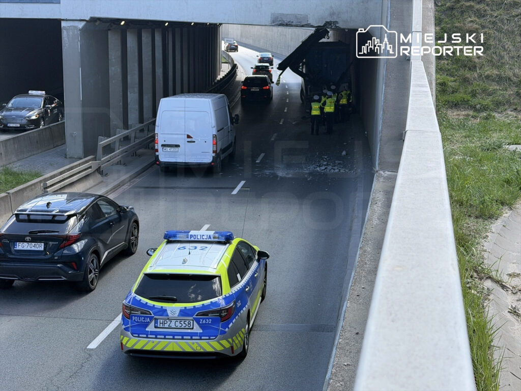 Grupa policjantów w kamizelkach odblaskowych bada uszkodzenie na dachu ciężarówki pod wiaduktem.