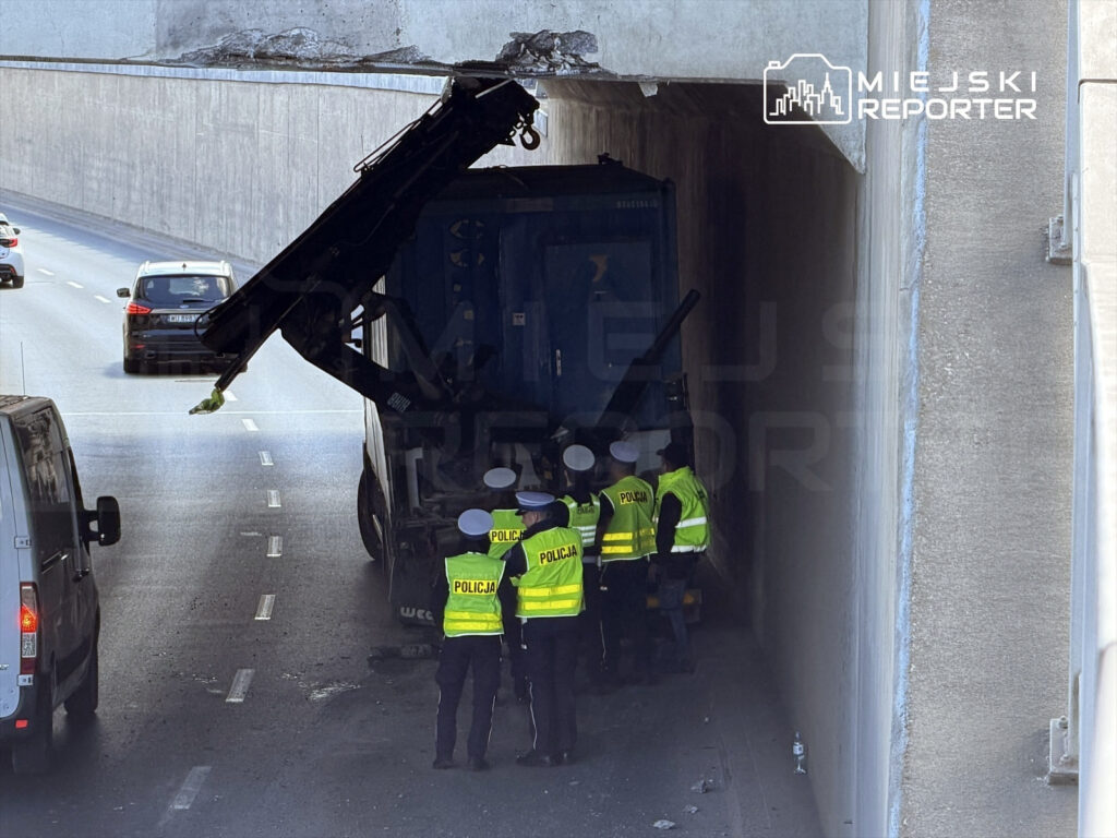 Grupa policjantów w kamizelkach odblaskowych bada sytuację przy unieruchomionej ciężarówce pod wiaduktem.
