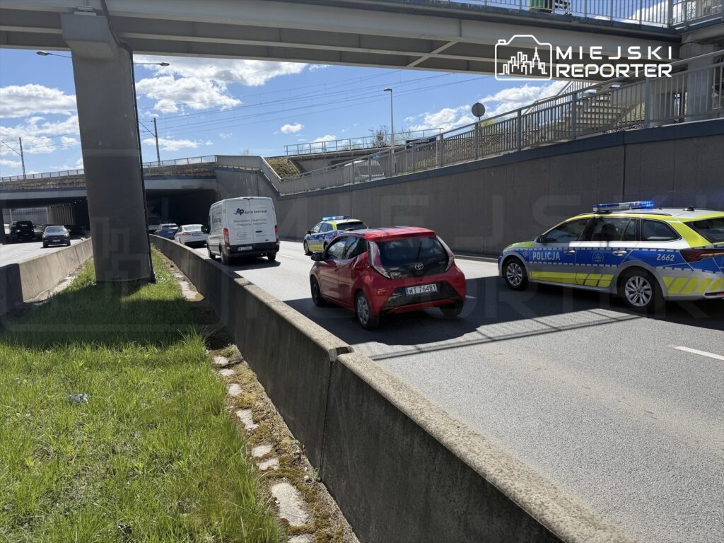 Ruch samochodowy na drodze z zatrzymanym pojazdem i radiowozem policji obok, w tle widoczne niebo i most.