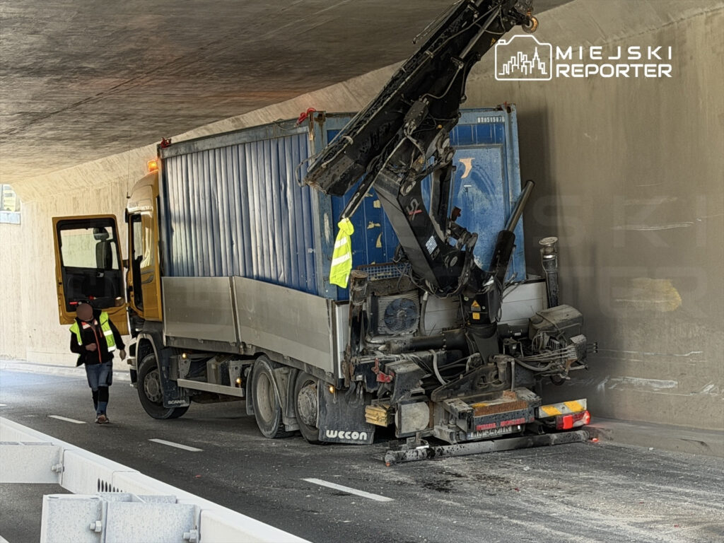 Ciężarówka z maszyną budowlaną pracuje w tunelu, a pracownik w kamizelce odblaskowej kontroluje postęp prac.