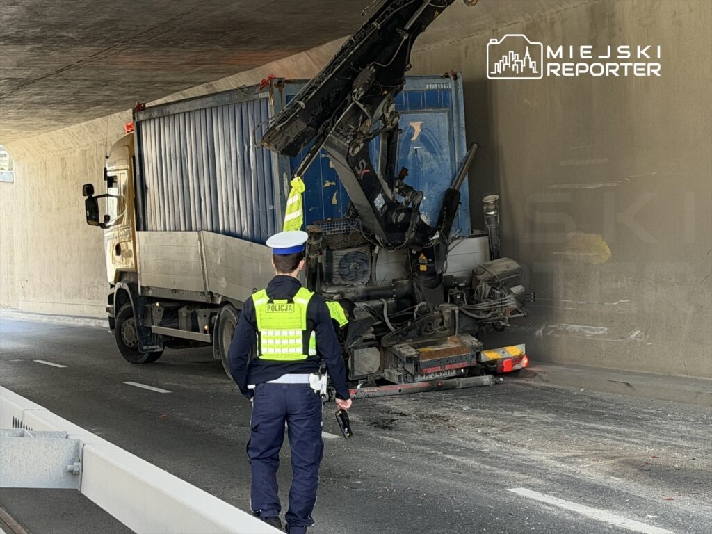 Kierowca ciężarówki z podniesioną naczepą utknął pod tunelem, a policjant kontroluje sytuację.