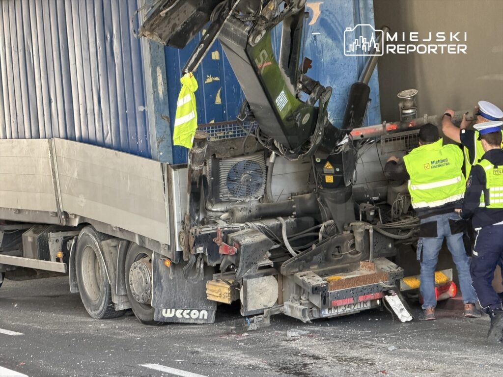 Wielki ciężarówka z uszkodzoną naczepą, obok niej pracują policjanci i pracownicy służb drogowych.