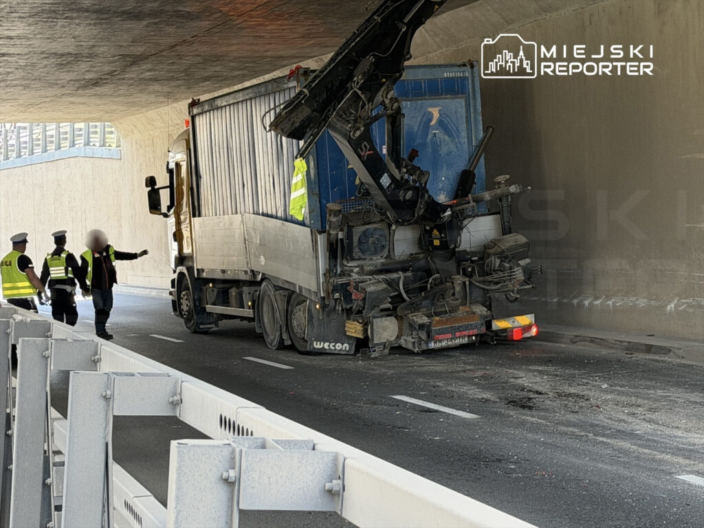 Trzech policjantów rozmawia z kierowcą ciężarówki, która utknęła pod wiaduktem. W tle widoczna uszkodzona naczepa.