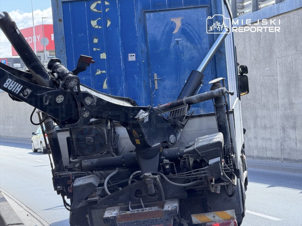 Czarny pojazd ciężarowy z podniesionym dźwigiem stoi na pasie ruchu obok szarego muru.