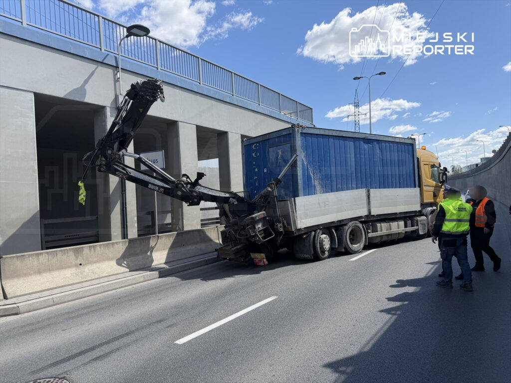 Ciężarówka z uniesionym dźwigiem utknęła pod wiaduktem, a pracownicy w kamizelkach odblaskowych oceniają sytuację.