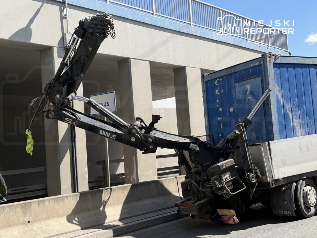 Wciągarka zamontowana na ciężarówce unosi niebieski kontener pod zadaszeniem parkingu.