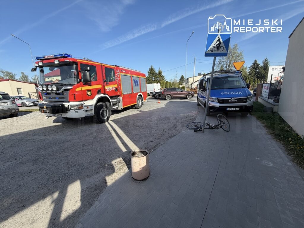 Czerwony wóz strażacki i niebieski radiowóz policyjny stoją na ulicy obok przewróconego roweru.