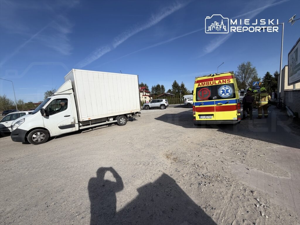 Kierowca białego samochodu ciężarowego zatrzymany na drodze, obok karetka i strażacy rozmawiający przy pojeździe.
