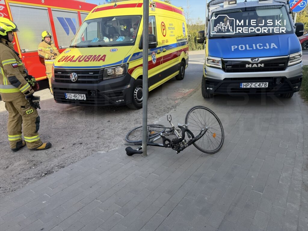 Strażak w mundurze obserwuje miejsce zdarzenia, obok leży przewrócony rower, a w tle stoją karetka i radiowóz policyjny.