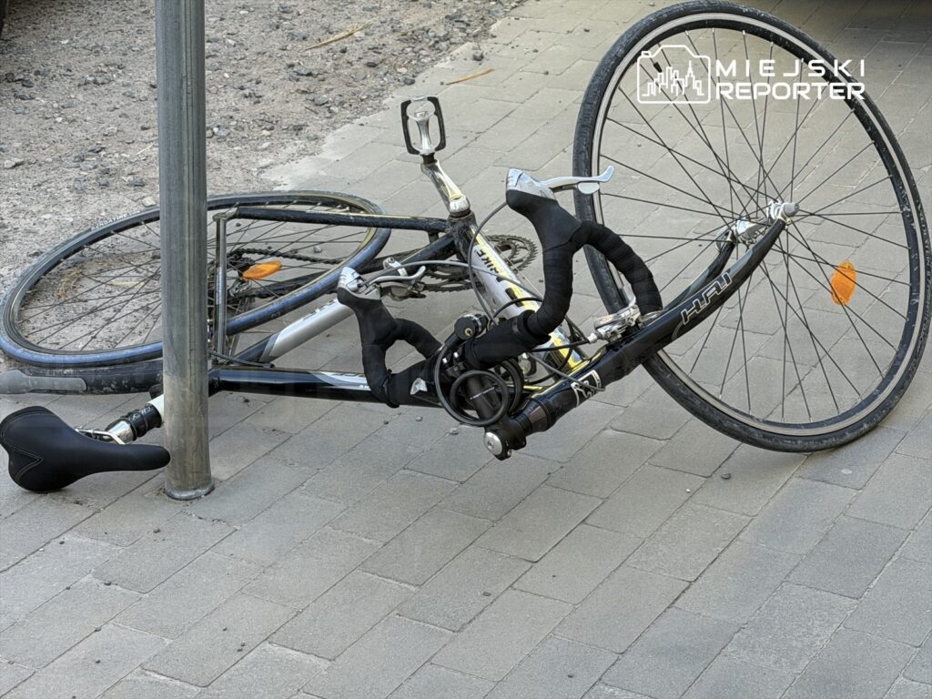 Złamany rower leżący na chodniku, przymocowany do słupa, z uszkodzoną przednią częścią i odwróconym kołem.