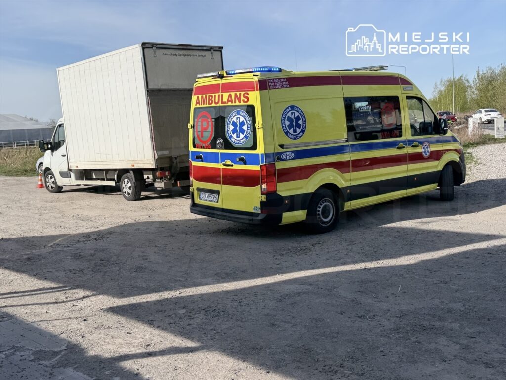 Karetka pogotowia parkowana obok białej ciężarówki na placu w otoczeniu zieleni i budynków.