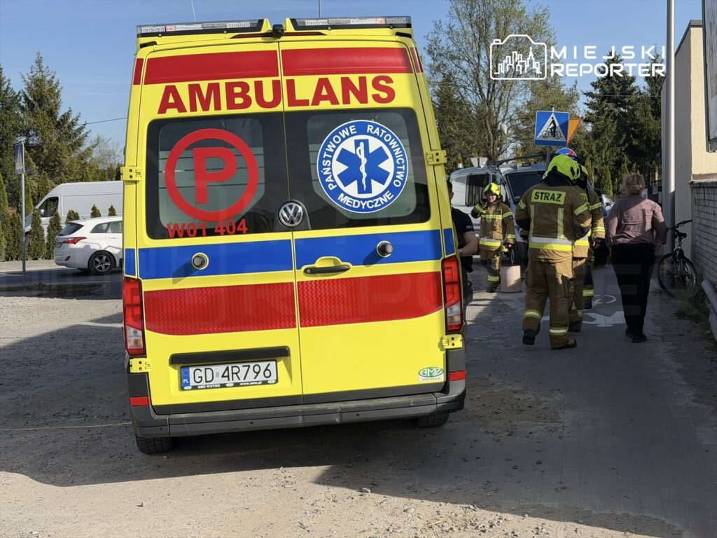 Zespół ratowników medycznych i strażaków pracuje obok karetki na ulicy, w tle widać zaparkowane samochody.