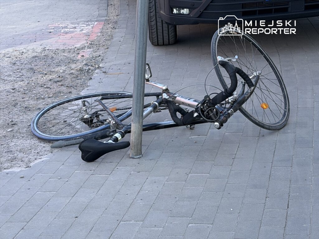 Przewrócony rower leży na chodniku obok metalowego słupa, w tle widoczna droga i opona pojazdu.