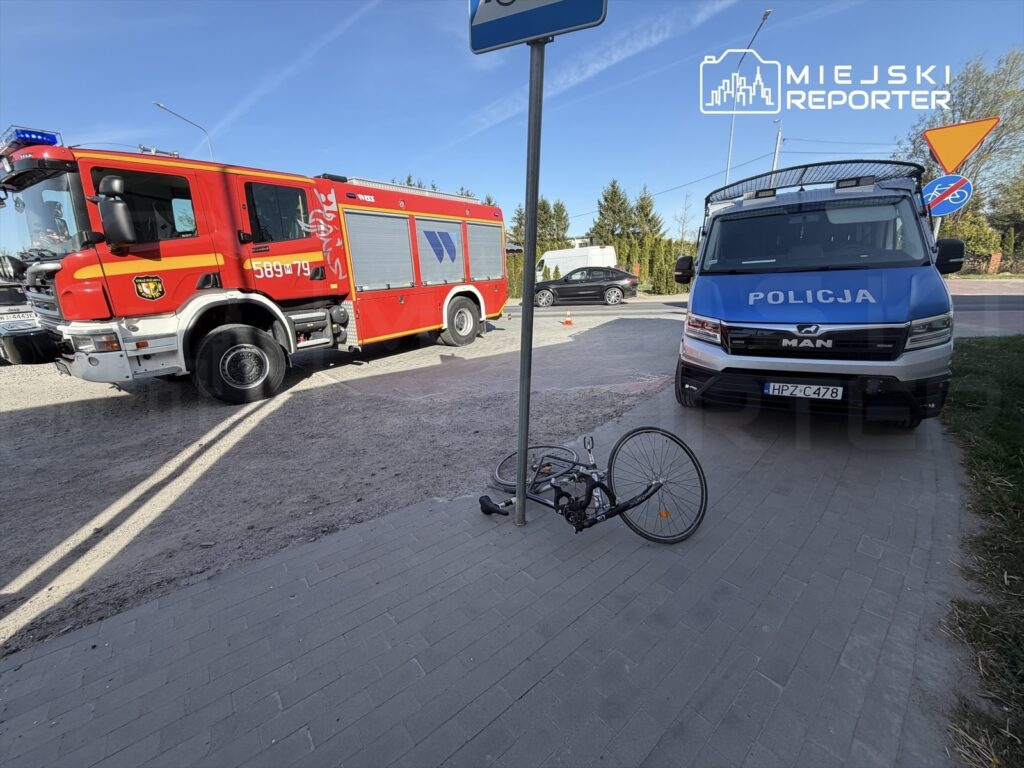 Czerwony wóz strażacki i niebieski radiowóz policyjny stoją obok przewróconego roweru na chodniku.