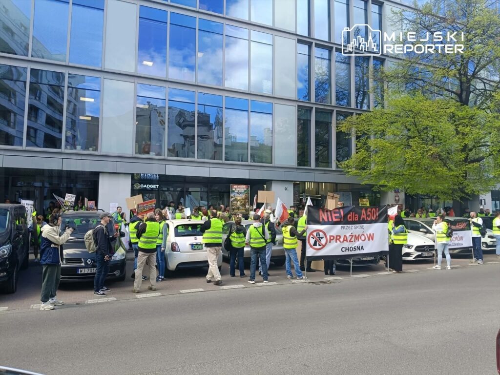 Grupa protestujących w żółtych kamizelkach trzyma transparenty z hasłami przed nowoczesnym biurowcem.