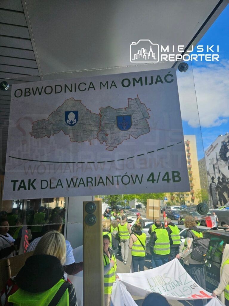 Grupa osób w kamizelkach odblaskowych trzyma transparenty i protestuje na ulicy, domagając się poparcia dla wariantu drogi.