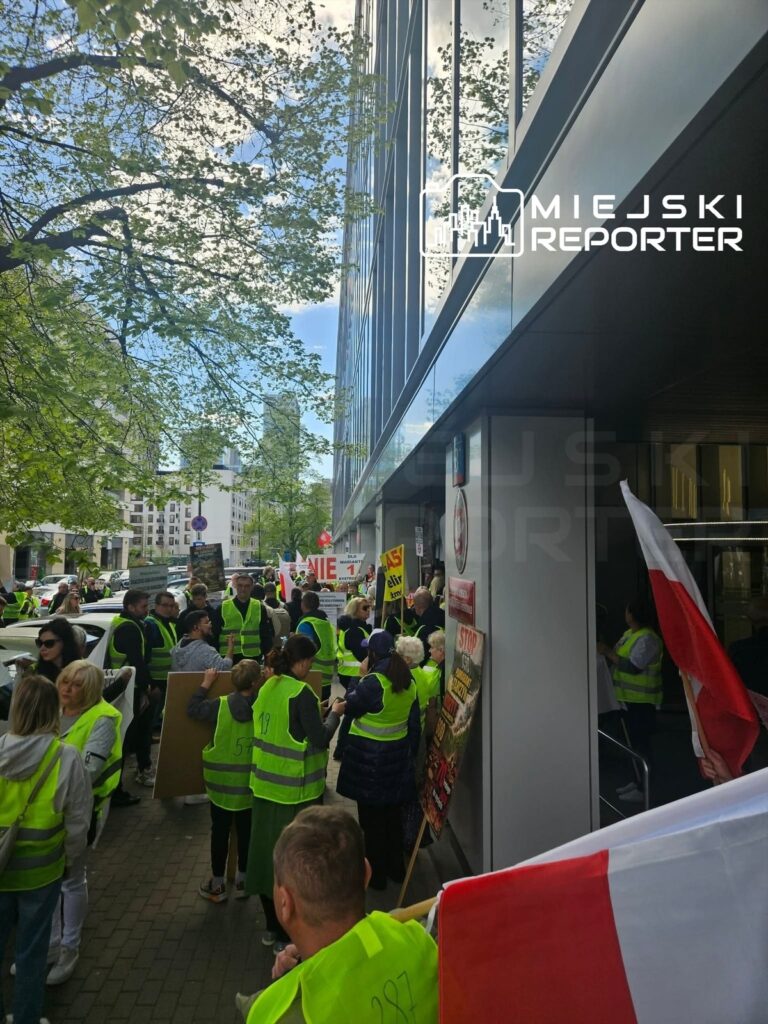 Grupa protestujących w kamizelkach odblaskowych z transparentami zbiera się przed nowoczesnym biurowcem.