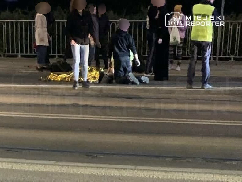 Grupa osób zgromadzona na ulicy, niektórzy pomagają osobie leżącej na jezdni, obok leży złota folia ratunkowa.