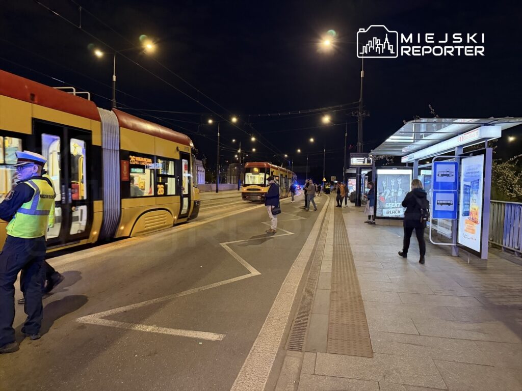 Mężczyzna w mundurze stoi obok tramwaju na przystanku, a pasażerowie czekają na tramwaj w nocy.