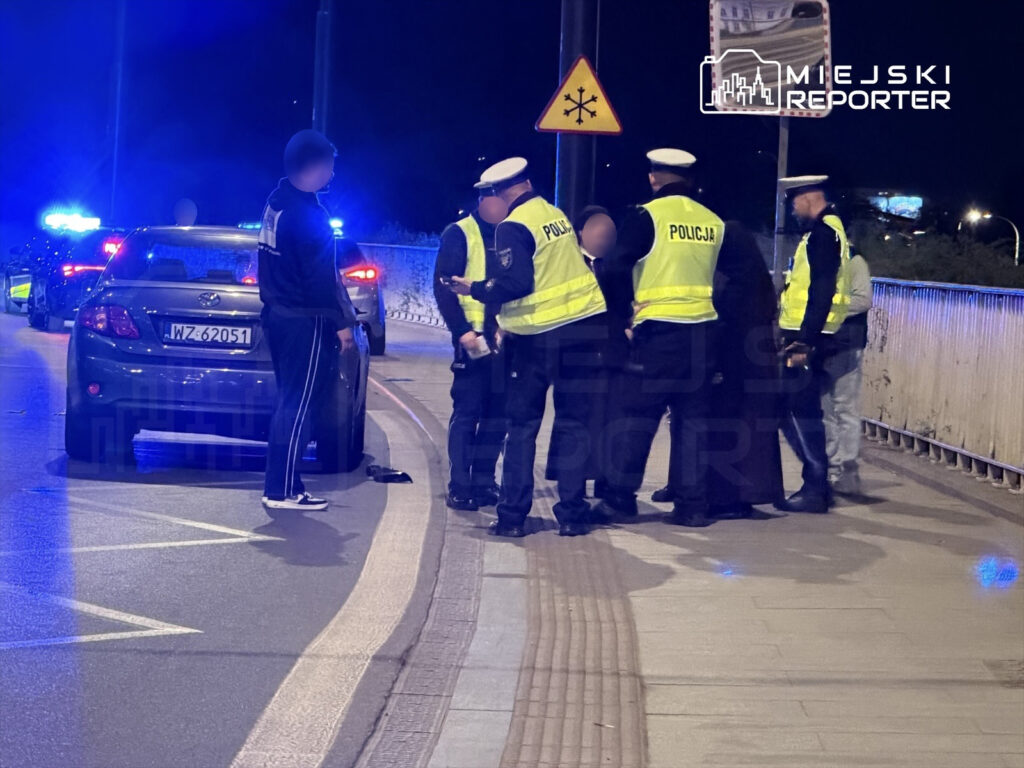 Policjanci w kamizelkach odblaskowych przeprowadzają interwencję w nocy przy zaparkowanym samochodzie osobowym.