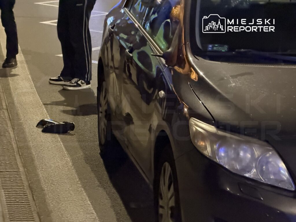 Czarna samochód zaparkowany na ulicy, obok leży uszkodzona część pojazdu. W tle widoczne nogi dwóch osób.