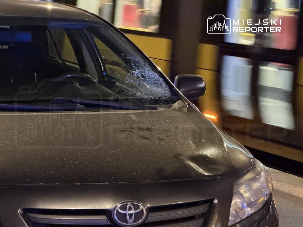 Czarny samochód osobowy z rozbitą szybą czołową stoi na ulicy obok przejeżdżającego tramwaju.