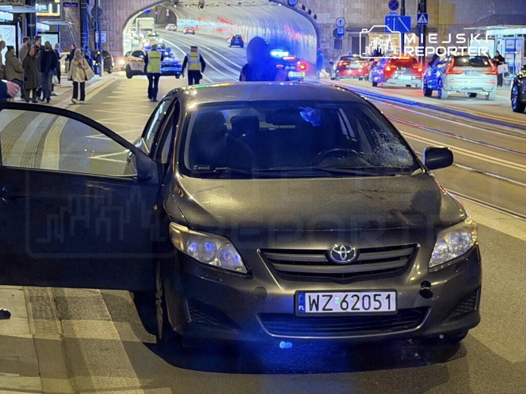 Czarny samochód osobowy z otwartymi drzwiami stoi na ulicy, obok policyjne radiowozy oraz przechodnie.