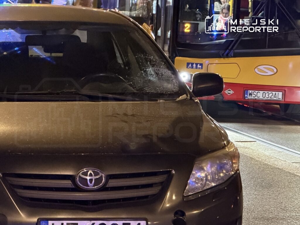 Uszkodzony samochód osobowy z pękniętą szybą stoi obok przystanku autobusowego, w tle widać autobus.