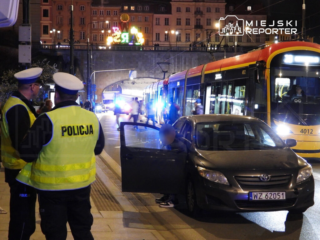Dwaj policjanci w odblaskowych kamizelkach rozmawiają obok zatrzymanego samochodu, w tle tramwaj i oświetlona ulica.