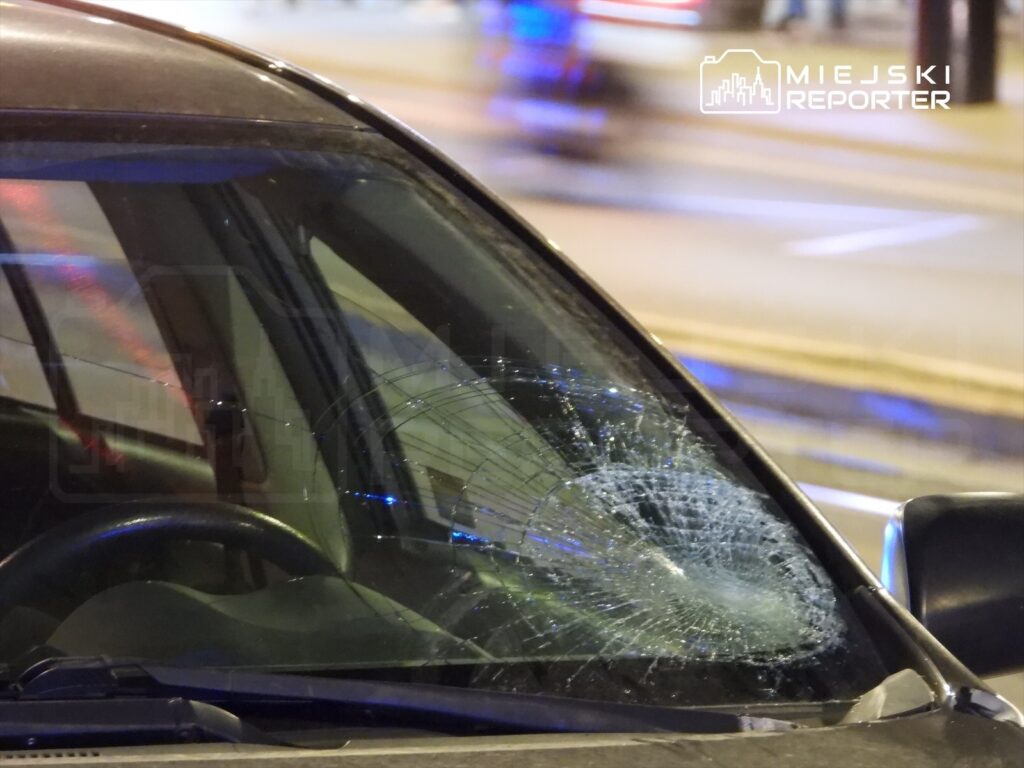 Pęknięta szyba czołowa samochodu stojącego na ulicy, w tle migające światła policyjnych radiowozów.