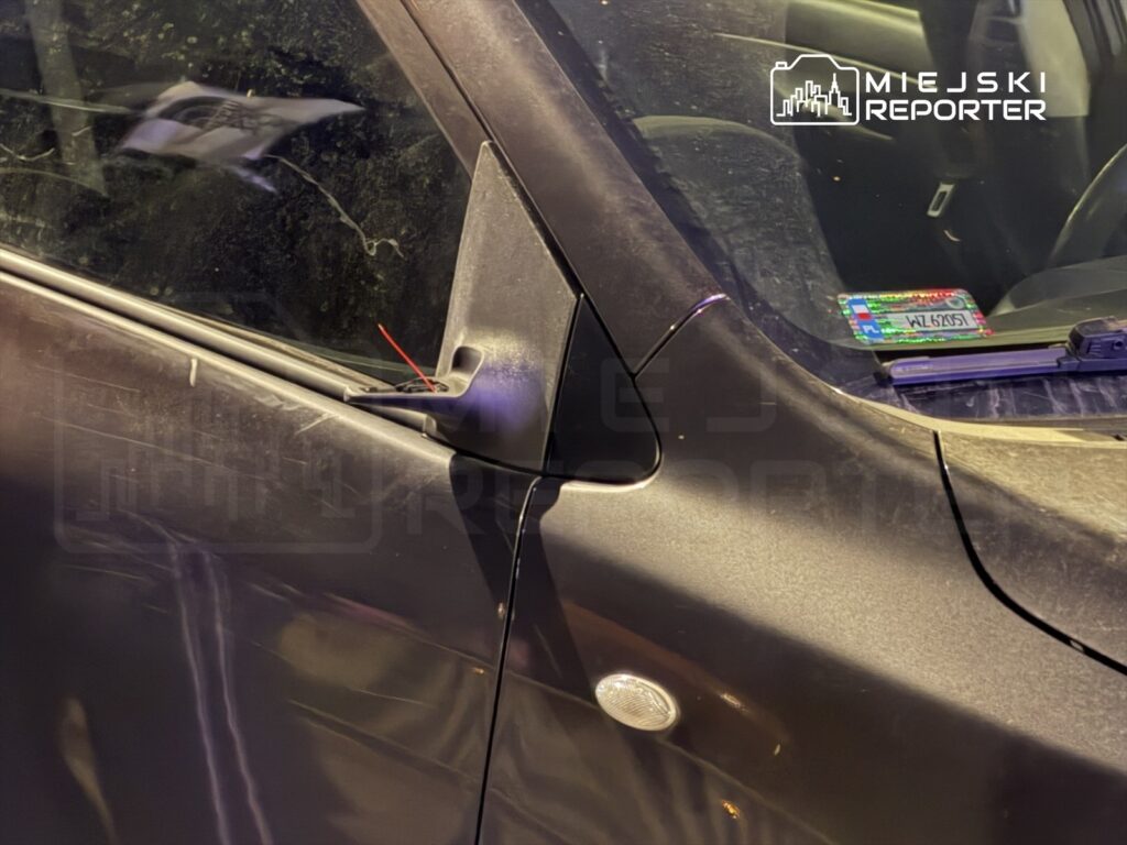 Czarny samochód osobowy z uszkodzoną boczną lusterkiem, stojący na parkingu z widoczną nalepką rejestracyjną na szybie.