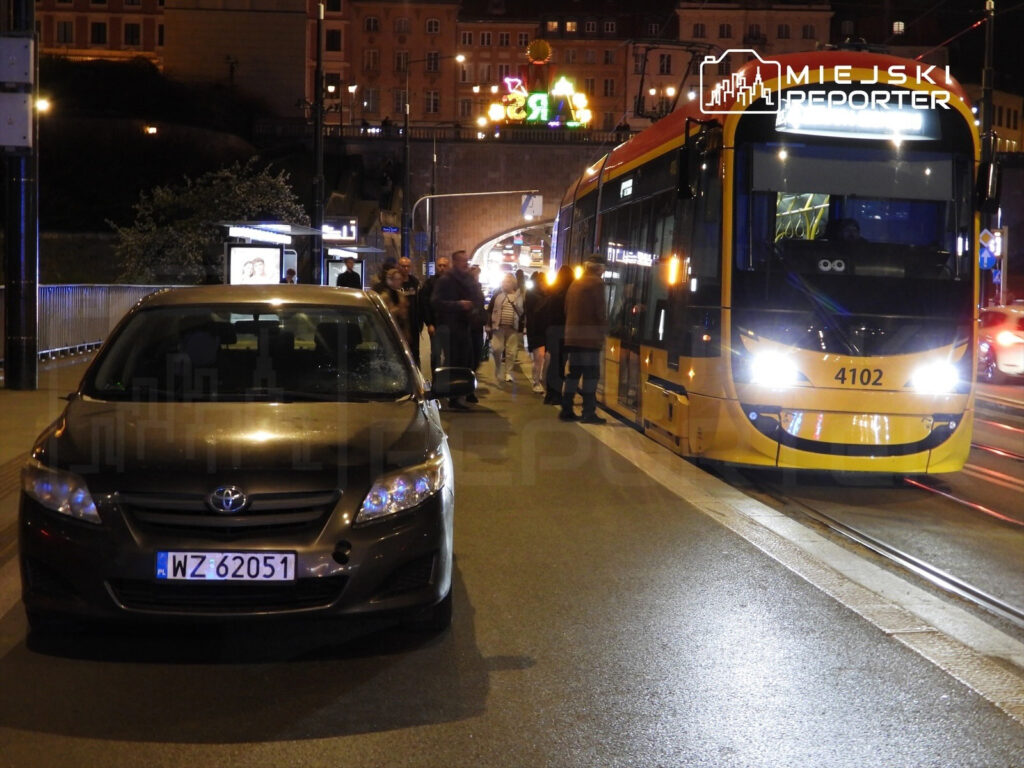 Czarny samochód osobowy zaparkowany na oświetlonej ulicy, obok tramwaju oraz grupy ludzi czekających na przystanku.