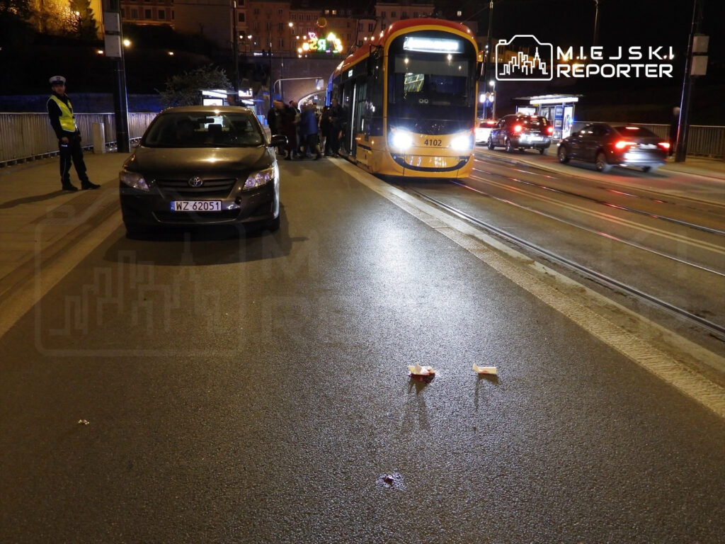 Na jezdni stoi samochód osobowy, obok grupka osób czeka na tramwaj, a w tle widać radiowoz policji. Na drodze leżą dwa opakowania.