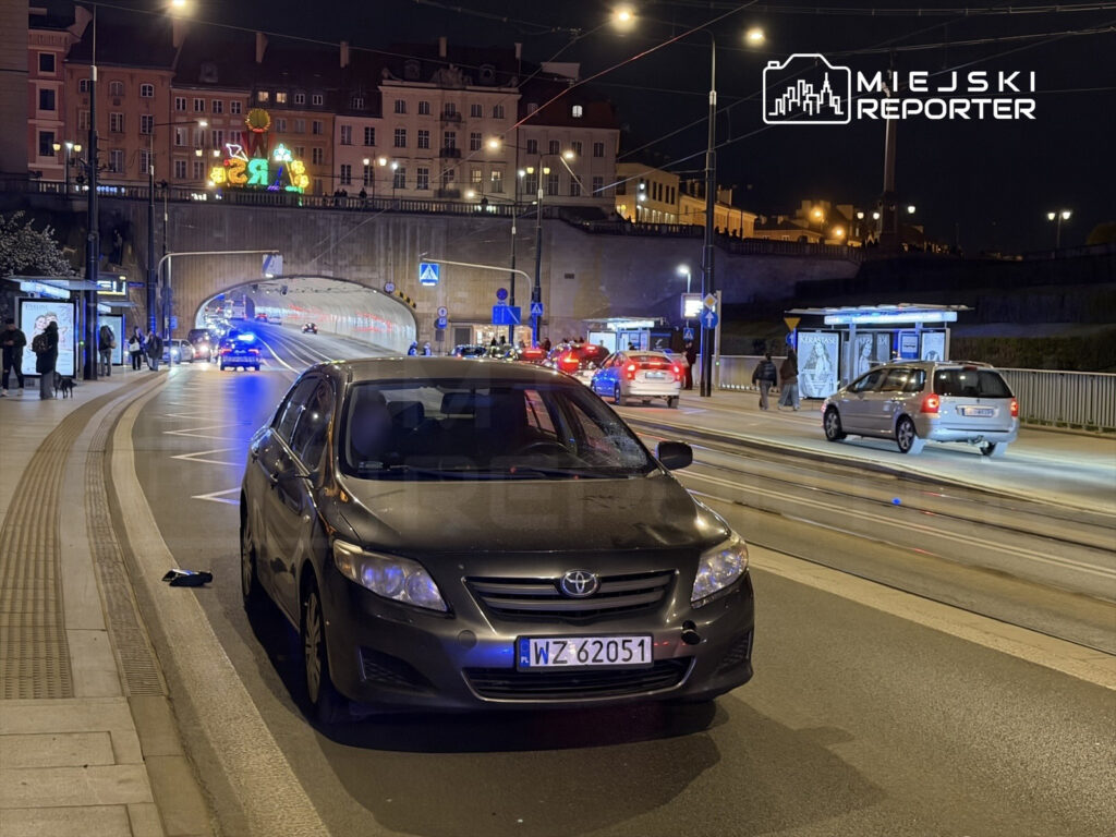 Samochód osobowy zaparkowany na ulicy w nocy, w tle widoczny tunel oraz oświetlone budynki.