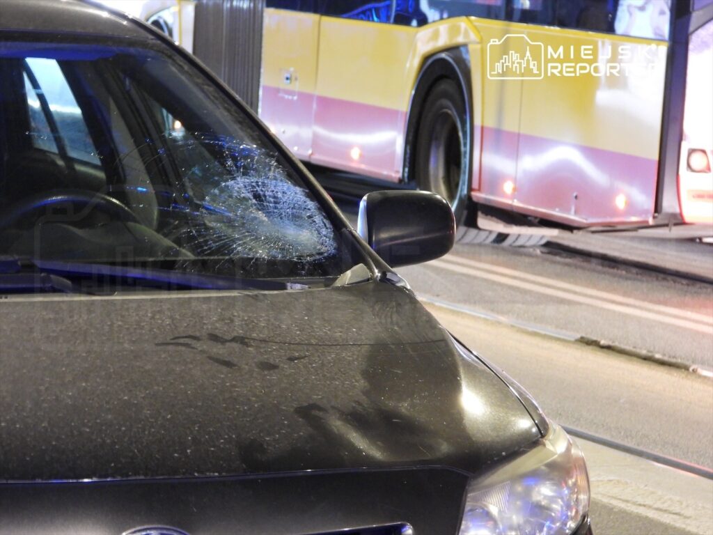 Uszkodzony samochód osobowy z pękniętą szybą przednią stoi na drodze obok przystanku autobusowego.