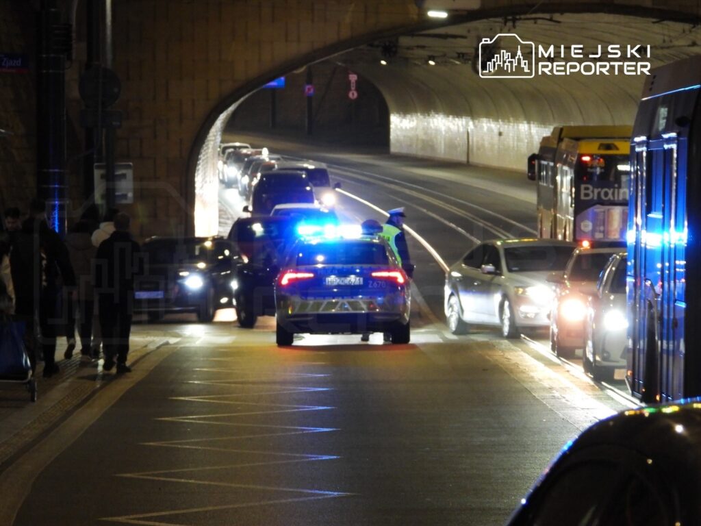Radiowóz policyjny z włączonymi sygnałami świetlnymi zatrzymuje ruch w tunelu, obok grupy pieszych.