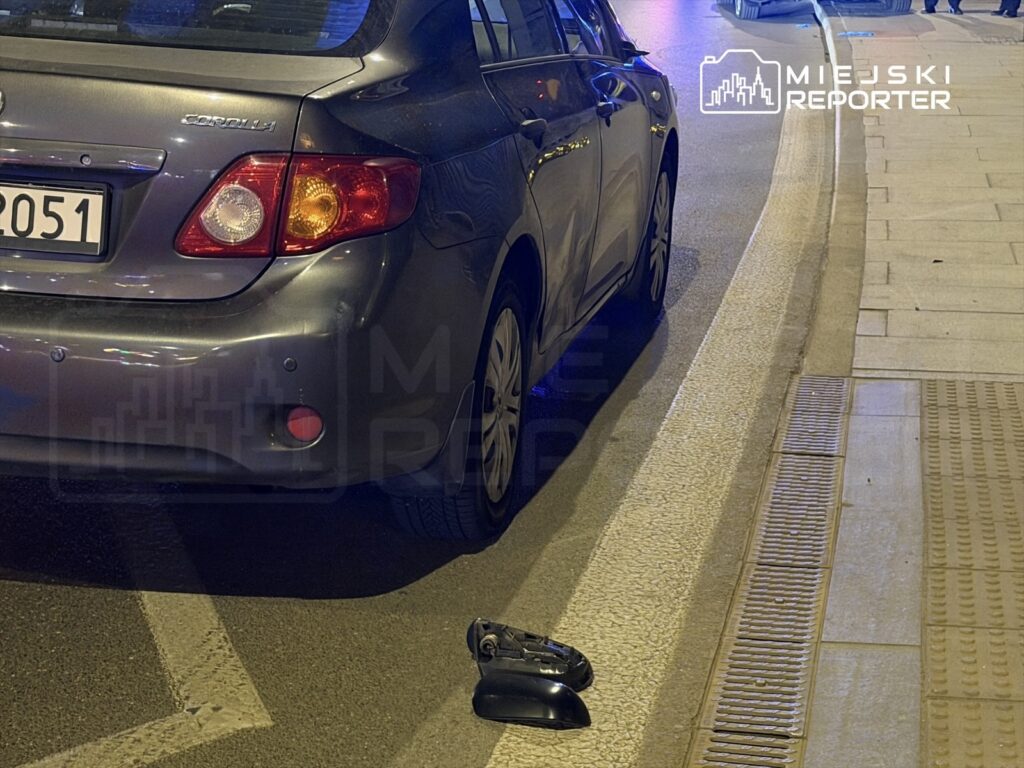 Szary samochód osobowy zaparkowany na ulicy, obok leży odłamany lusterko.