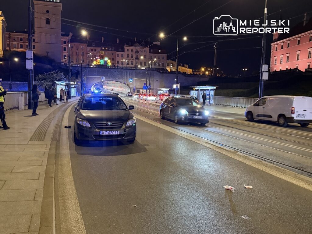 Czarny samochód osobowy zaparkowany na ulicy w nocy, obok grupy ludzi i oznakowanej stacji tramwajowej.