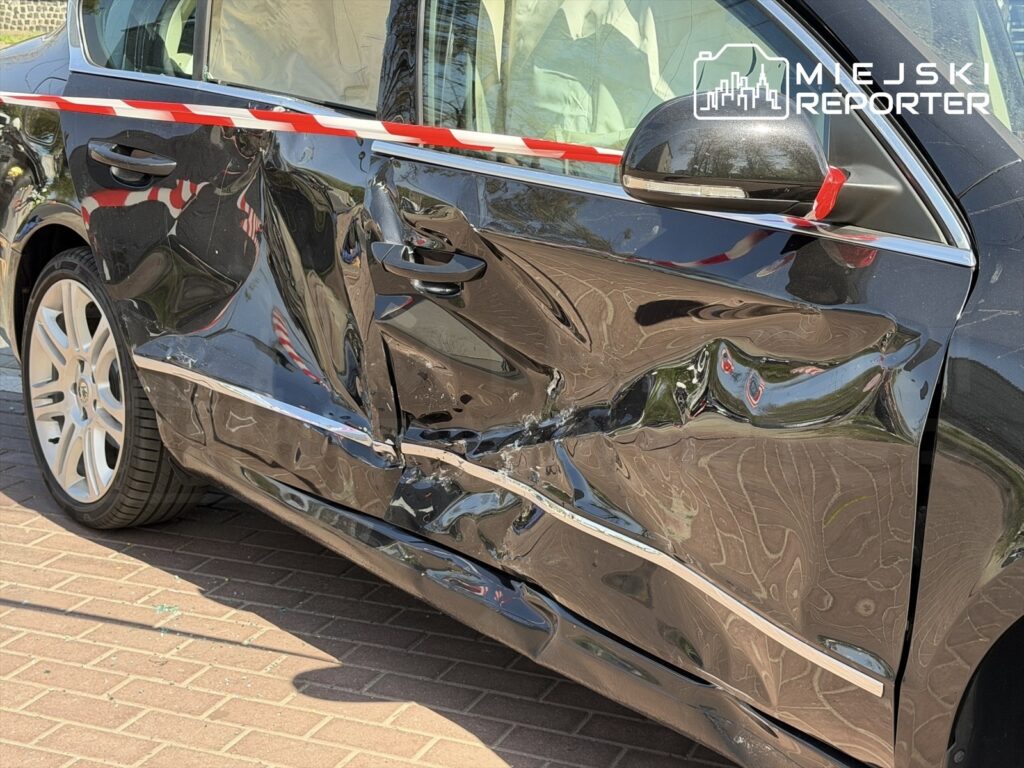 Uszkodzony czarny samochód osobowy z wgnieceniem na drzwiach, zaparkowany na brukowanej nawierzchni.