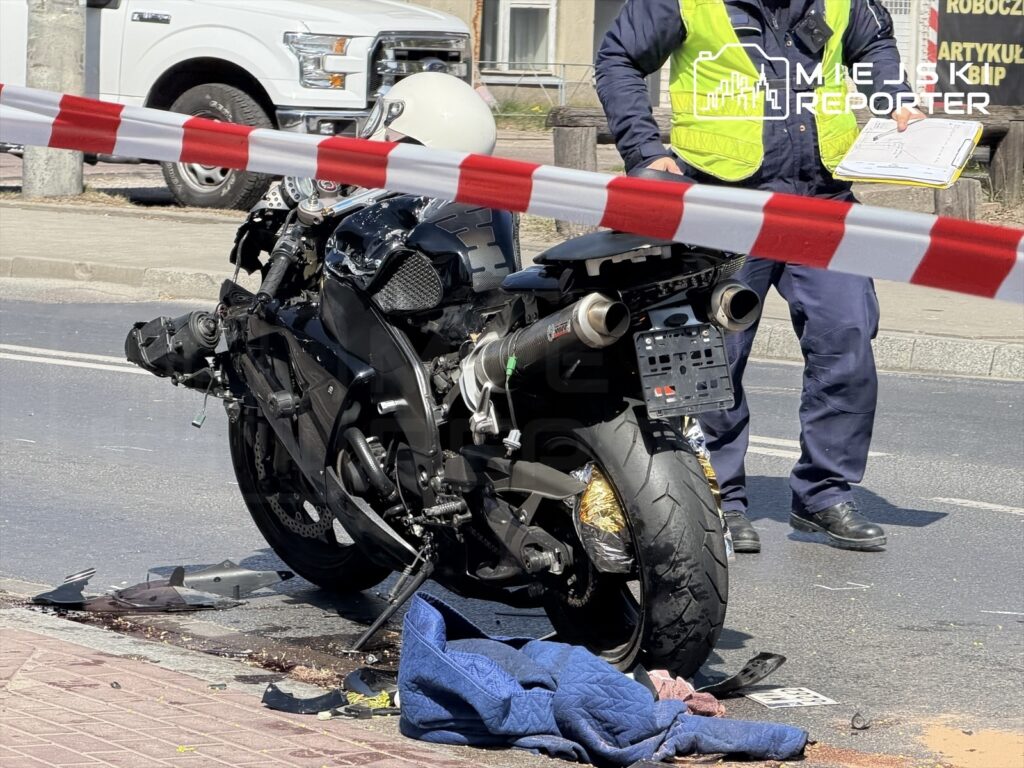 Motocykl leży na jezdni obok policjanta w kamizelce odblaskowej, zabezpieczony taśmą. Na drodze widoczne ślady płynów.