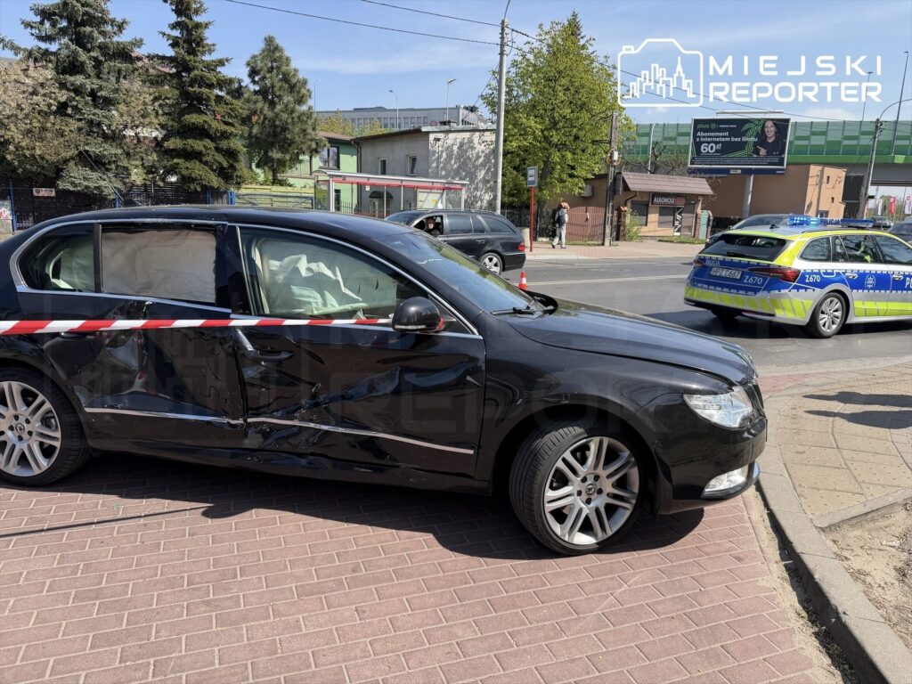Czarny samochód osobowy z uszkodzoną lewą stroną stoi na chodniku, otoczony taśmą zabezpieczającą. W tle widać radiowóz policyjny.