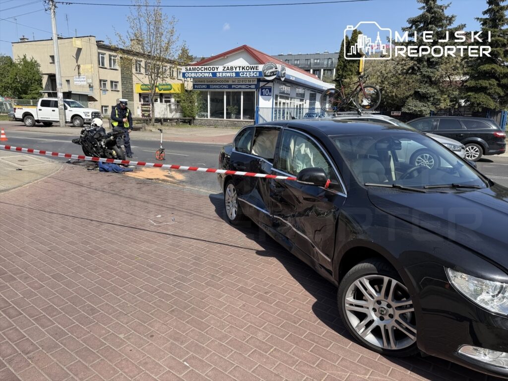 Motocyklista leży obok przewróconego motocykla, a policjant zabezpiecza miejsce zdarzenia taśmą. Na ulicy widoczne są uszkodzone pojazdy.