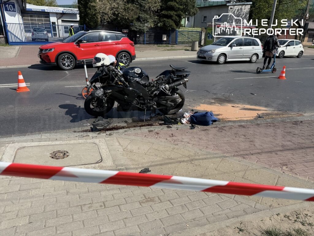 Motocykl leży na drodze obok rozlanej substancji, a w tle widać zaparkowane samochody i funkcjonariuszy w kamizelkach.