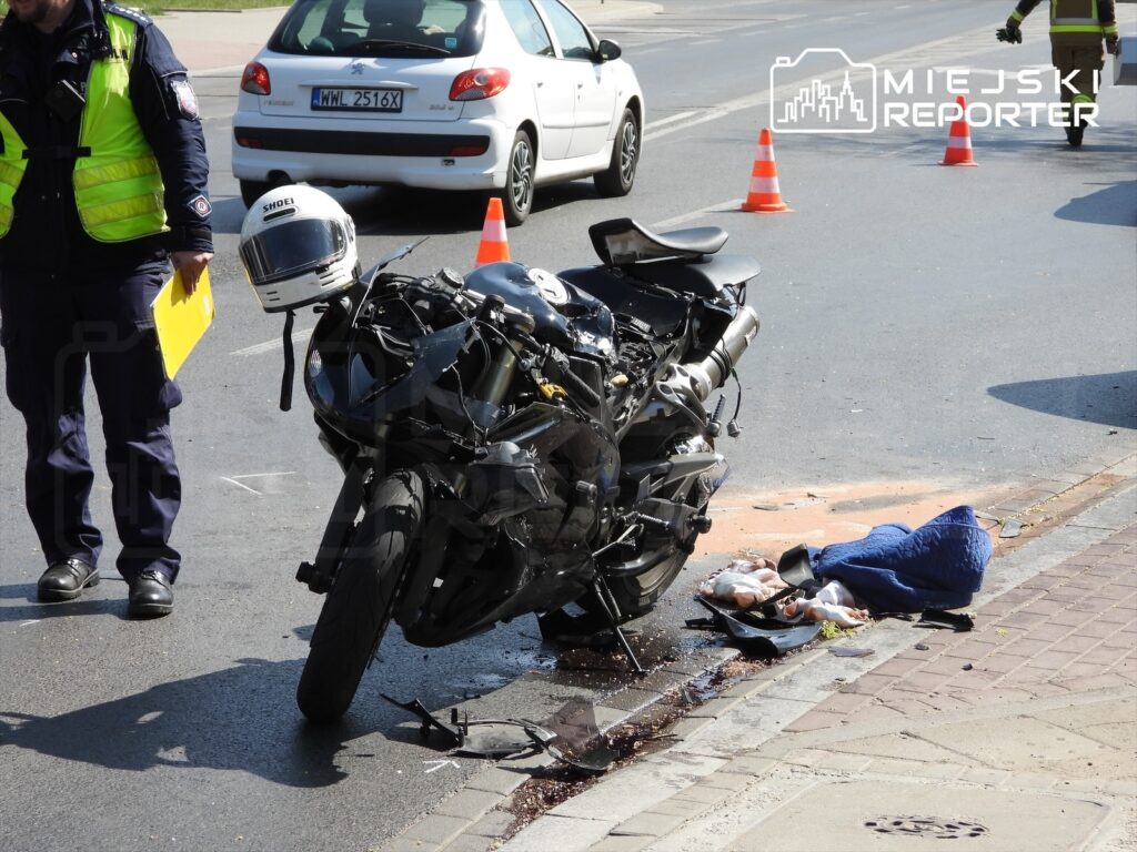 Motocykl leżący na boku na jezdni, obok niego policjant z notatnikiem, w tle zaparkowane samochody i pachołki.
