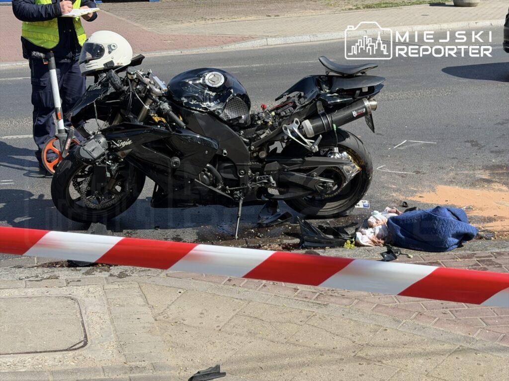 Motocykl po wypadku leży na jezdni, obok stoi policjant z notatnikiem, a teren jest zabezpieczony taśmą.