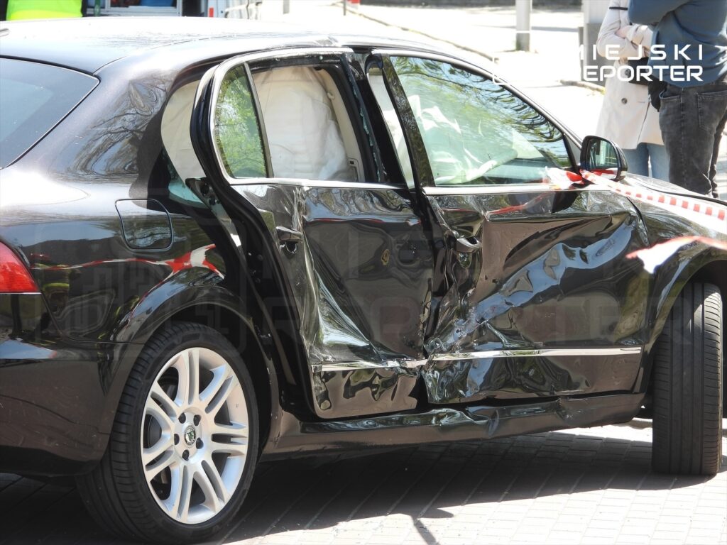 Czarny samochód osobowy z uszkodzoną lewą stroną stoi na chodniku, obok widoczni świadkowie zdarzenia.