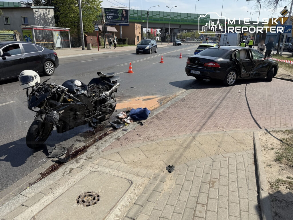Motocykl leży na poboczu z uszkodzeniami, obok znajduje się czarny samochód z wgniecioną stroną. Na jezdni widoczna krew.