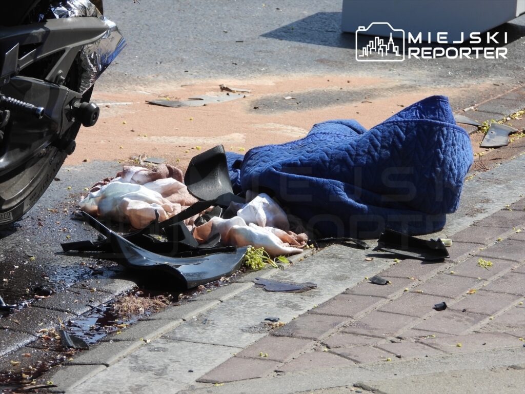 Zniszczona motocyklowa owiewka leży na chodniku obok plamy płynów i porozrzucanych materiałów.